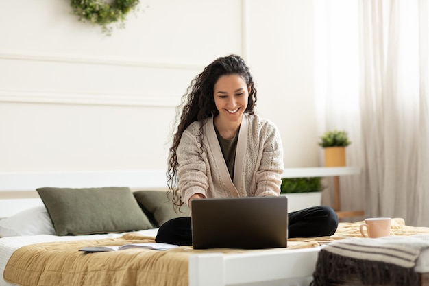 Radosna młoda dama siedzi ze skrzyżowanymi nogami na łóżku i używa laptopa do pracy online lub edukacji w domu kopia