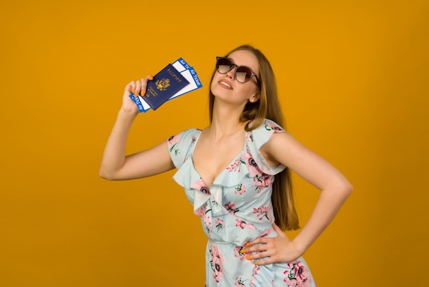 Radosna młoda kobieta w niebieskiej sukience z kwiatami i okularami przeciwsłonecznymi trzyma bilety lotnicze z paszportem na żółtym tle. Cieszy się z wznowienia turystyki po pandemii koronowirusa.