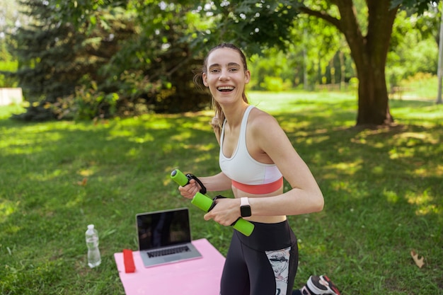 Radosna pani trzyma hantle stojąc na kolanach przy laptopie w zielonym letnim parku