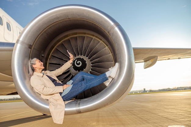 Radosna podróżniczka robiąca zdjęcie smartfonem i uśmiechająca się podczas odpoczynku w silniku turbiny samolotu