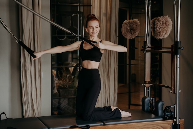 Radosna, szczupła, ruda kobieta na reformatorze pilates wykonująca ćwiczenia rozciągające ramiona podczas treningów w studio fitness, okazująca pewność siebie w każdym ruchu, wysportowana kobieta w stroju sportowym