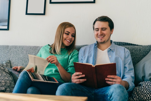 Radosna uśmiechnięta młoda kobieta i mężczyzna w zwykłych ubraniach, odpoczywający na przytulnej szarej kanapie podczas czytania książki