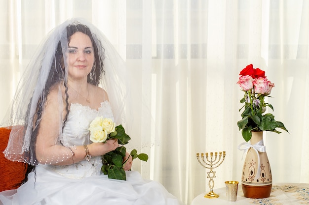 Radosna żydowska panna młoda z twarzą zakrytą welonem z bukietem białych róż siedzi w synagodze przed odprawieniem ceremonii Huppa przy stole z kwiatami