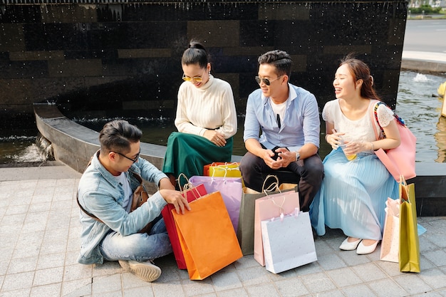 Radosni przyjaciele siedzą na zewnątrz z papierowymi torbami po zakupach w centrum handlowym