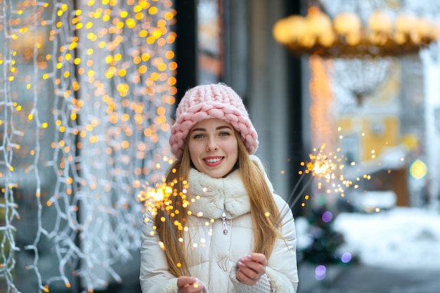 Radosny blond kobieta nosi dzianinowy kapelusz i ciepły płaszcz trzymając ognie. Świąteczny strzał na zewnątrz