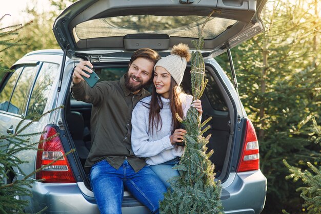 Radosny Brodaty Mężczyzna I ładna Kobieta W Kapeluszu Robiącym Selfie Zdjęcie Siedzi W Bagażniku Samochodu Z Jodły Na Terenie Plantacji.