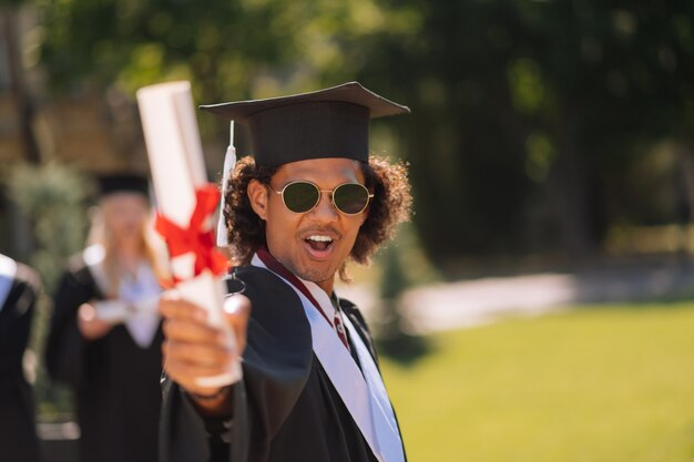 radosny chłopiec stojący przed swoją uczelnią z dyplomem w ręku