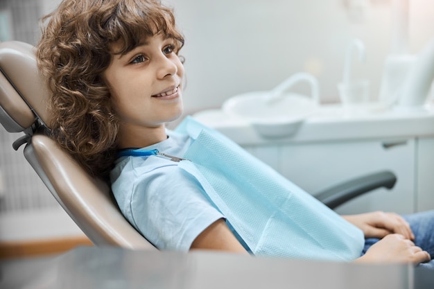 Radosny dzieciak z kręconymi włosami uśmiechający się siedząc na fotelu dentystycznym podczas wizyty u dentysty