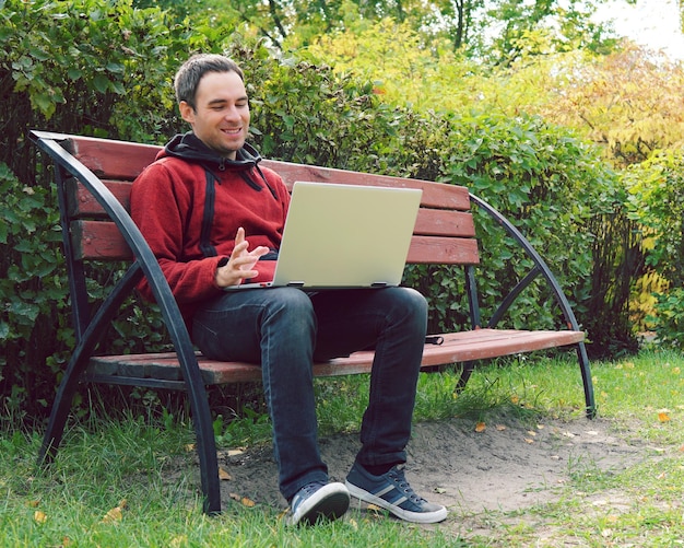 Radosny facet komunikuje się przez Internet na ulicy Programista korzysta z notebooka siedząc w jesiennym parku Przystojny mężczyzna pracuje na swoim laptopie Bezpłatna praca na zewnątrz freelancer na zewnątrz