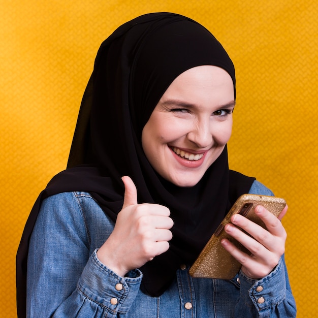 Radosny kobiety mienia telefon komórkowy gestykuluje thumbup przeciw żółtej powierzchni