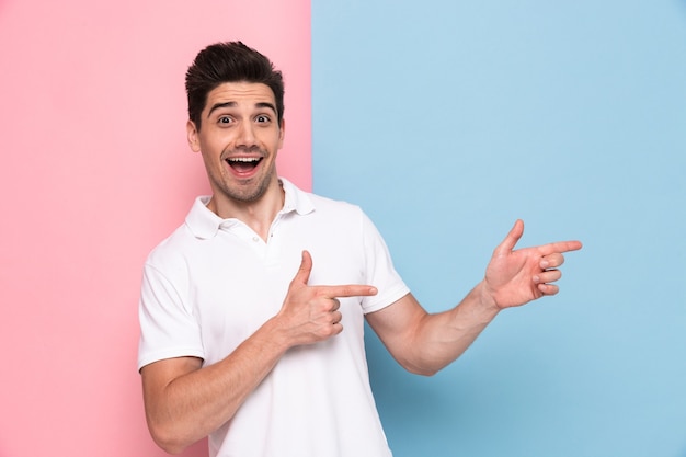 Radosny mężczyzna w casual t-shirt uśmiechnięty i pokazujący copyspace na dłoni, na białym tle nad kolorową ścianą