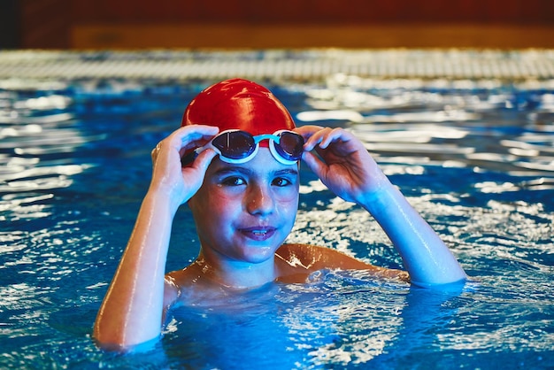 Radosny uśmiechnięty chłopiec pływak w czapce i goglach uczy się profesjonalnego pływania na basenie w siłowni z bliska