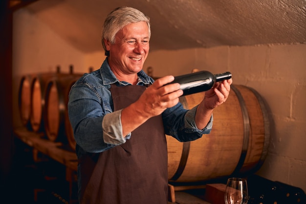 Radosny winiarz w fartuchu trzymający butelkę napoju alkoholowego i uśmiechnięty podczas spędzania czasu w przechowalni wina