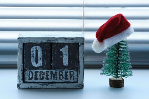 Ranek 01 grudnia na drewnianym kalendarzu stoi obok ozdobionej choinki w czerwonej czapce Świętego Mikołaja na oknie z żaluzjami