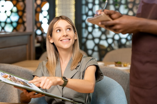 Ręce afrykańskiego kelnera stojącego przy stole i przyjmującego zamówienie blond uśmiechniętej dziewczyny wskazującej na menu podczas zamawiania deseru lub sałatki w kawiarni