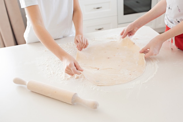 Ręce dzieci rozwałkują ciasto do pizzy na białym stole. Wspólna zabawa w kuchni. Widok z góry.