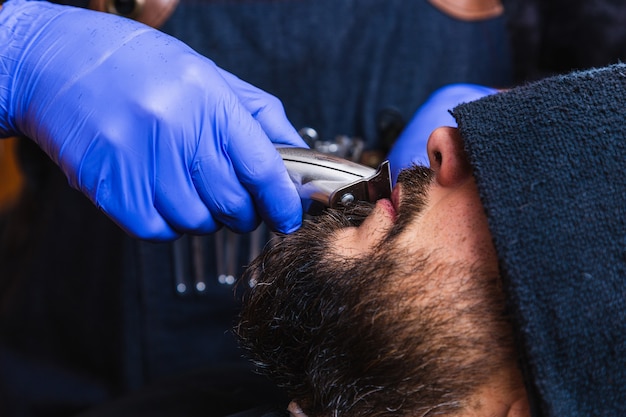 Ręce fryzjera obcinają brodę klientowi golarką elektryczną.