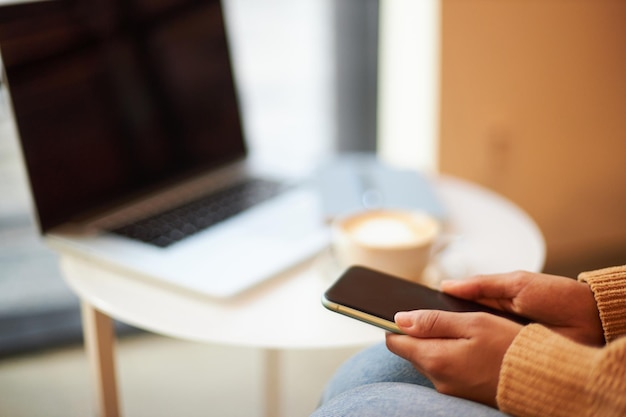 Ręce kobiety trzymającej smartfon z pustym ekranem siedzącej przy stole z laptopem i filiżanką kawy