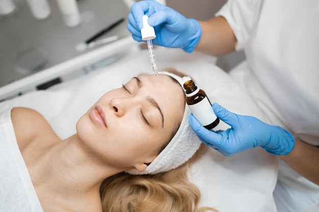 Ręce kosmetologa ociekające odżywczym olejem skórę twarzy młodej kobiety w salonie spa