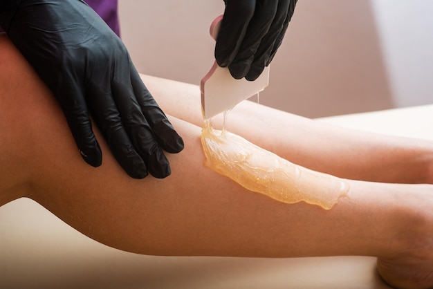 Zdjęcie ręce kosmetologa w czarnych rękawiczkach nakładających pastę cukrową szpachelką na nogę kobiety procedura depilacji