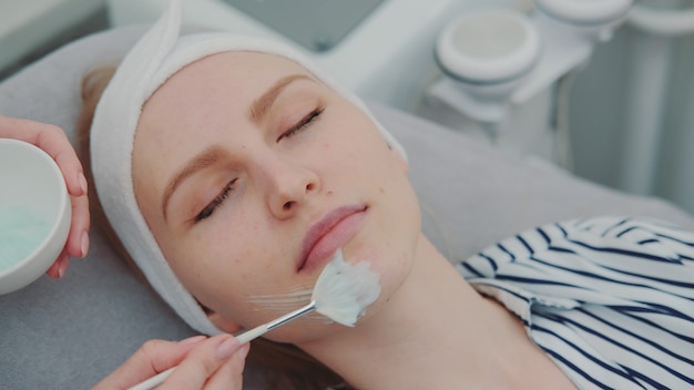 Ręce kosmetyczki stosowania maski krem na twarz młodej kobiety w salonie spa