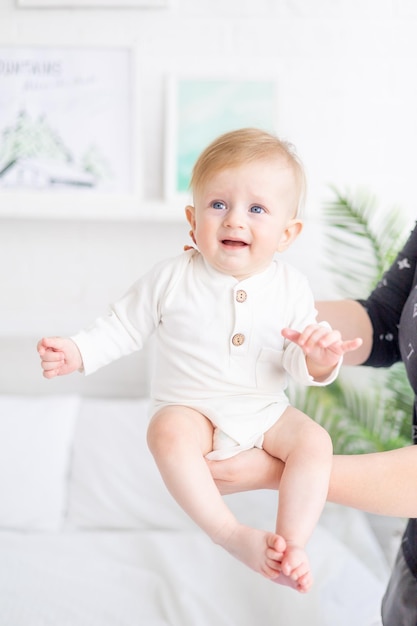 Ręce mamy trzymają niemowlę w wieku 6 miesięcy blond chłopiec o niebieskich oczach na białym łóżku w jasnej sypialni w białym bawełnianym body koncepcja towarów dla dzieci