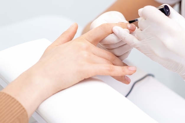 Ręce manikiurzystki pokrywają bezbarwnym lakierem do paznokci kobiety w salonie manicure.