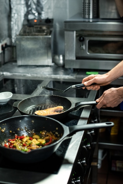 Ręce męskiego szefa kuchni pieczenie kawałka łososia na gorącej patelni z oliwą z oliwek, stojąc przy kuchence elektrycznej i gotując ryby i warzywa