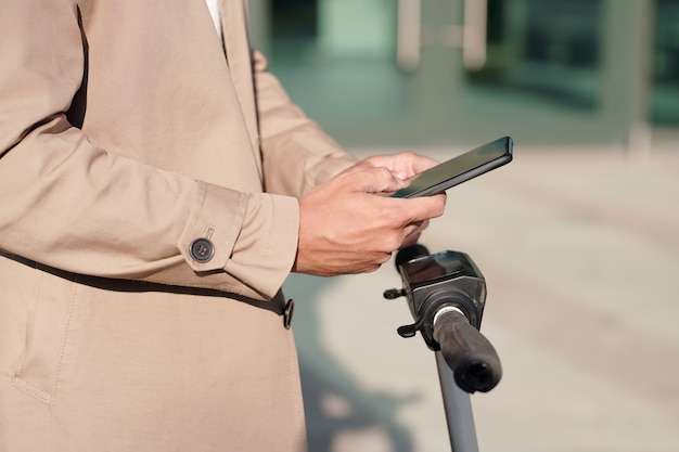 Ręce młodego biznesmena mobilnego w beżowym płaszczu wiadomości w smartfonie na uchwytach skutera elektrycznego w środowisku miejskim