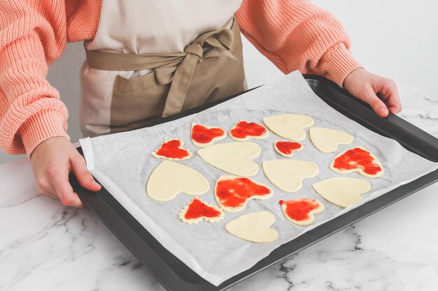 Ręce młodej białej piekarki trzymają arkusz do pieczenia z sercowymi ciasteczkami posypanymi surowymi