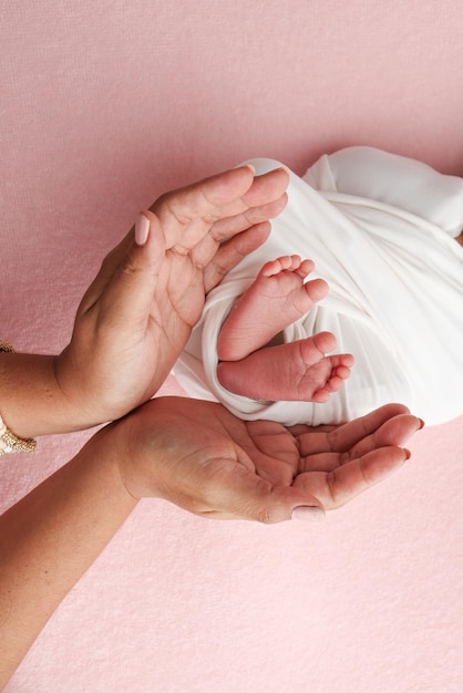 Zdjęcie ręce ojca, matka trzymają stopę noworodka w białym kocyku na różowym tle, nogi noworodka na dłoniach rodziców, zdjęcie palców dzieci, pięty i stopy.