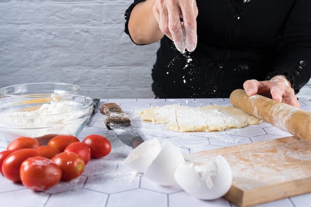 Ręce rzucające mąkę na rozciągnięte ciasto ze składnikami i narzędziami do gotowania
