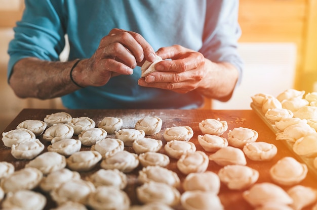 Ręce Starszego Mężczyzny, Gotowania I Formowania Małych Domowych Niegotowanych Pierogów Z Mięsem Na Kuchennym Stole