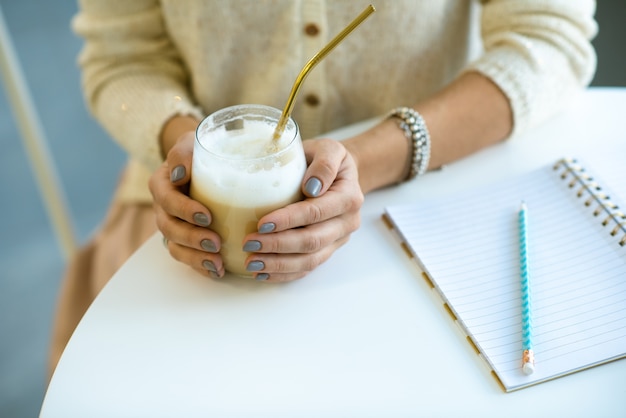 Ręce studentka trzymając szklankę cappuccino ze słomką, siedząc przy stole przy przerwie i odrabianiu lekcji
