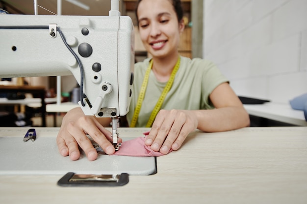 Ręce szczęśliwego młodego krawca pracującego na elektrycznej maszynie do szycia na poduszkach na ramiona dla nowej sukienki, płaszcza lub innego elementu odzieży