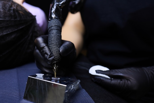 Ręce Tatuażysty W Czarnych Rękawiczkach, Trzymającego W Jednej Ręce Maszynkę Do Tatuażu, A W Drugiej Bawełnianą Gąbkę