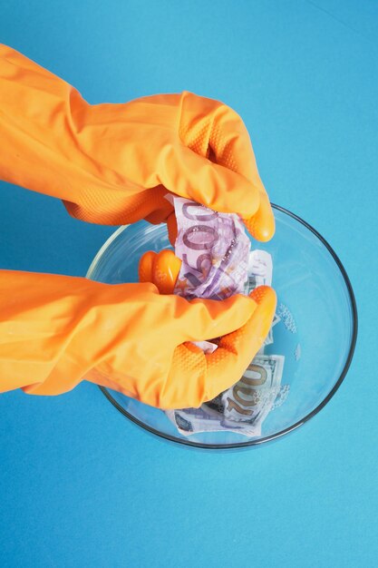Ręce w pomarańczowych rękawiczkach myją pieniądze mydłem na niebieskim tle
