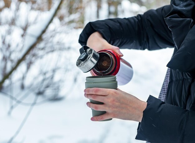 Ręce wlewające gorącą herbatę do kubka z termosu na zewnątrz podczas zimowych wakacji podczas leśnego spaceru