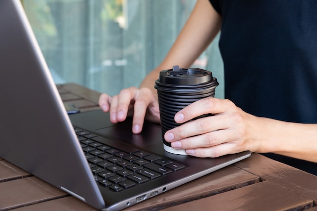 Ręce Womans z papierową filiżanką kawy lub herbaty i laptopa w kawiarni lub na tarasie domowym