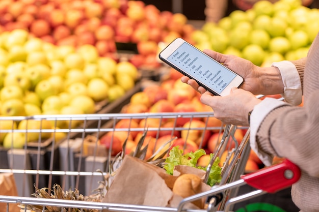 Ręce współczesnej dojrzałej klientki ze smartfonem nad produktami spożywczymi w koszyku przeglądając listę zakupów