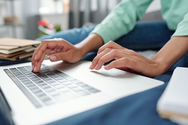 Ręce współczesnej studentki rasy mieszanej, naciskając przyciski klawiatury, siedząc na łóżku przed laptopem i korzystając z sieci