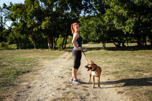 Redhair jouful młoda kobieta pieszcząca swojego psa w sportowej odzieży, ciesząc się swoim czasem i wakacjami w słonecznym parku