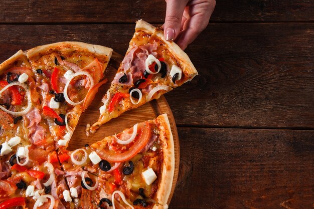 Ręka biorąc gorący kawałek kolorowe pyszne włoskie pizze serwowane na drewnianym stole, leżał płasko. Ciemne tło z miejsca na kopię.