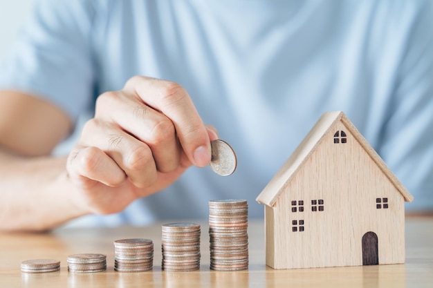Ręka człowieka umieścić monetę na stosie monet z drewnianym domem na stół z drewna. oszczędność pieniędzy na zakup domu, koncepcja kredytu mieszkaniowego planu finansowego.