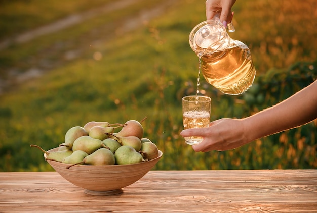 Ręka kobiety nalewa świeży smaczny sok gruszkowy do szkła z naturalnym tłem sadu na świetle zachodzącego słońca Skład owoców wegetariańskich