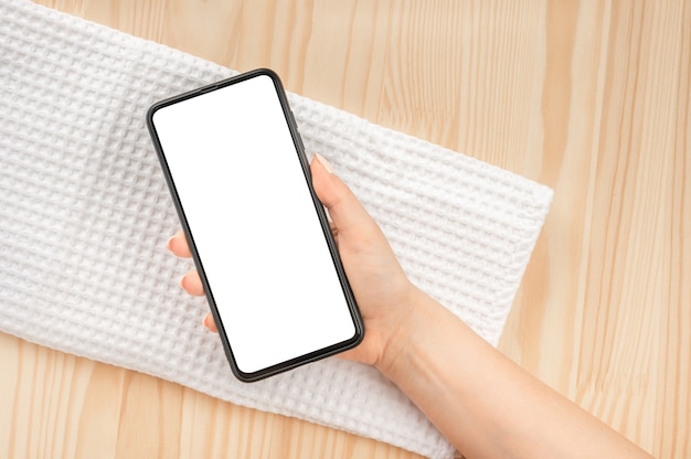 Ręka kobiety trzymać makieta telefon komórkowy z pustego ekranu na naturalny drewniany stół i biały wafel ręcznik do zabiegów spa. pusty biały ekran reklam spa i kosmetyków naturalnych