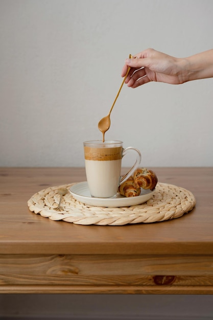 Ręka kobiety z łyżką nad filiżanką kawy z mlekiem i rogalikami na drewnianym stole