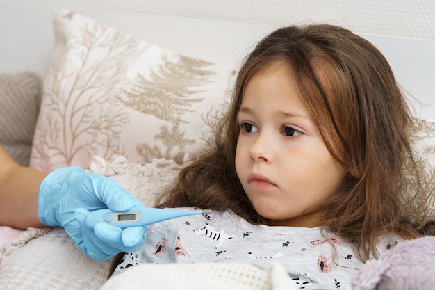 Ręka lekarza w rękawiczce medycznej mierzy temperaturę chorej dziewczynki za pomocą elektronicznego termometru Gorączka ciepła w łóżku