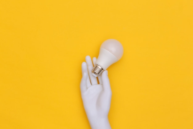 Ręka manekina białego trzyma żarówkę LED na żółtym tle. Widok z góry