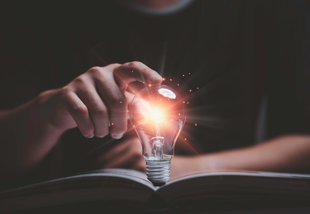 Ręka mężczyzny dotyka świecącej żarówki na otwartej książce do czytania, aby szukać wiedzy i koncepcji innowacyjnego pomysłu kreatywnego myślenia